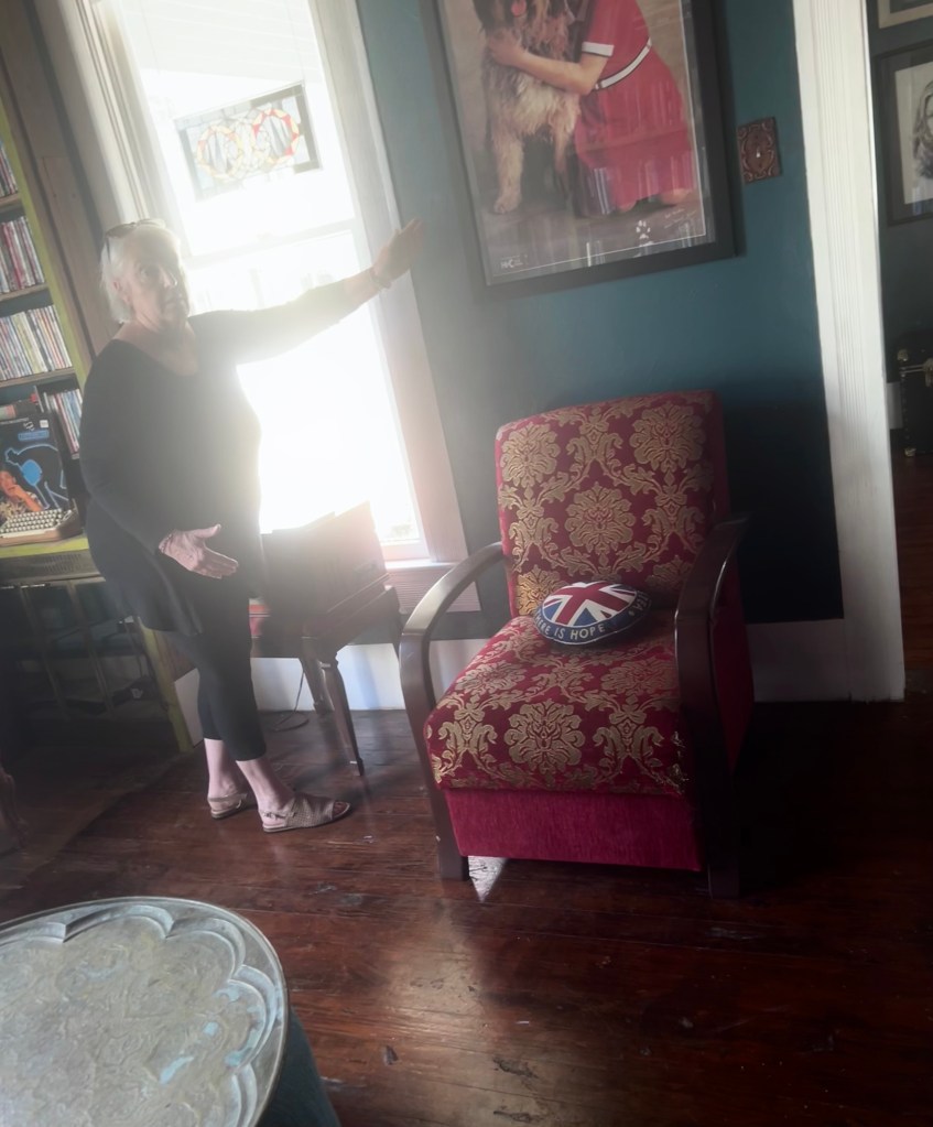 An older woman gesturing toward a window in a room with wooden floors, a red upholstered chair, and a decorative pillow featuring a British flag.
