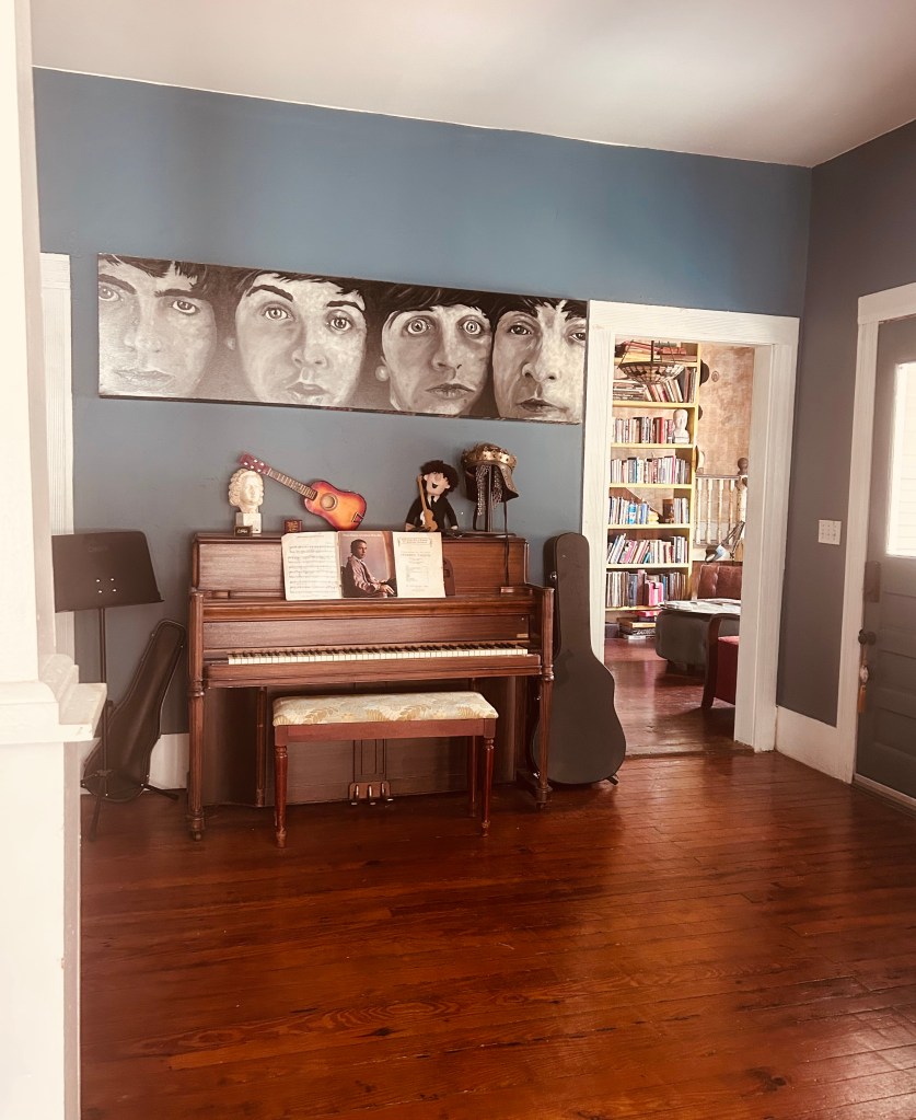 A cozy room featuring a wooden piano under a painting of four faces, surrounded by musical instruments and decor, with a doorway leading to a bookshelf area.